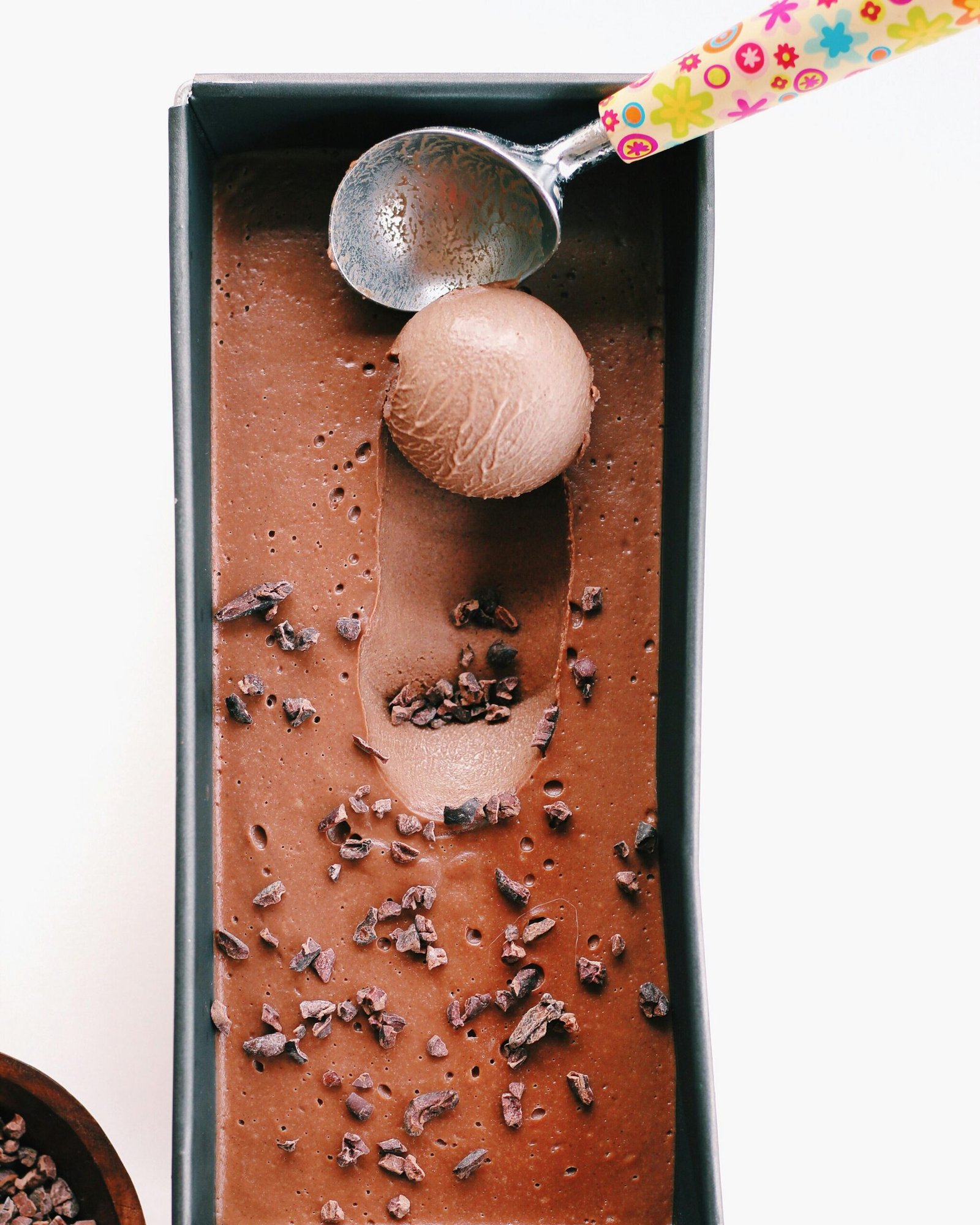 Delicious homemade chocolate ice cream in a tray with a colorful scoop, garnished with cacao nibs.
