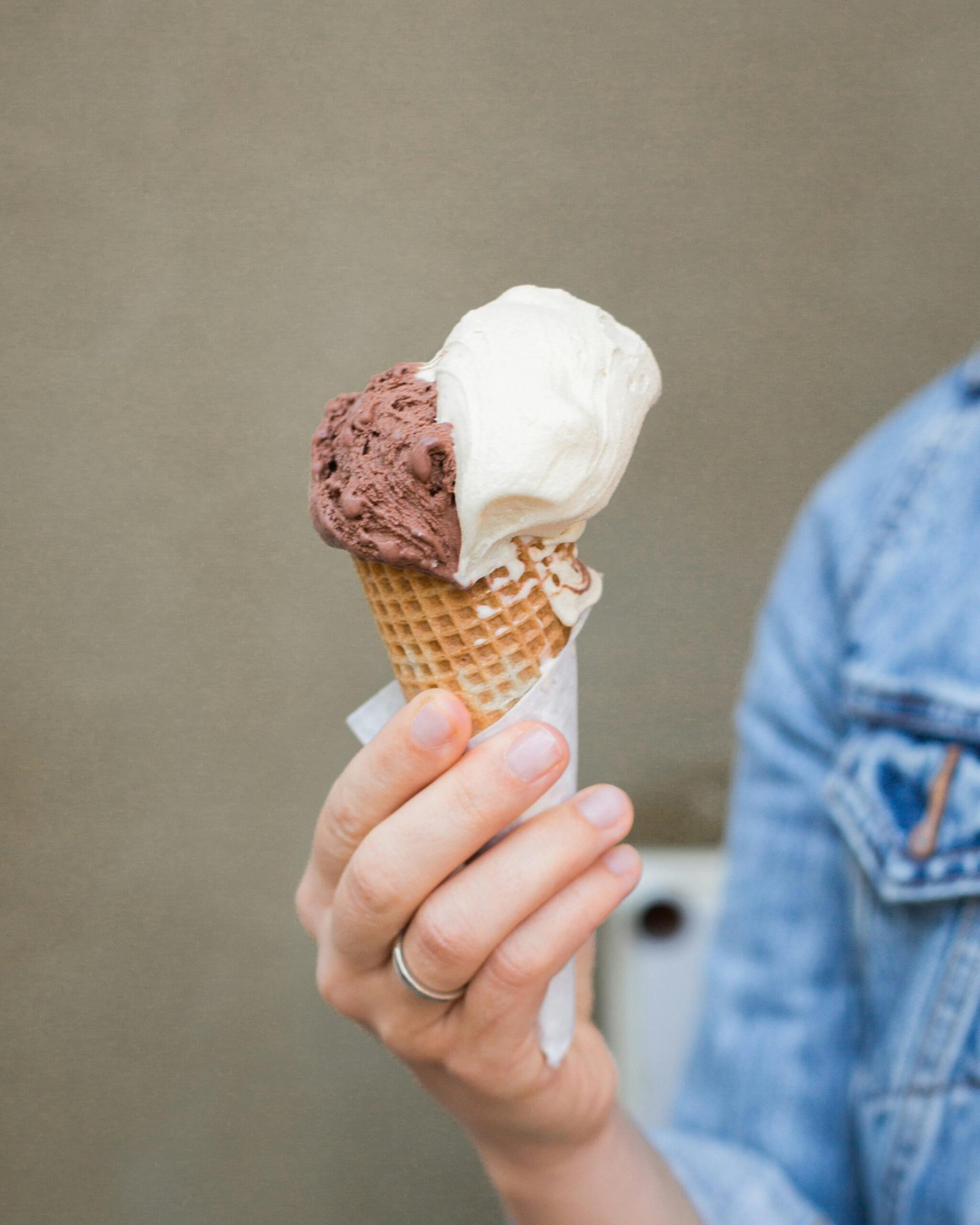 Chocolate and vanilla ice cream cone held by person in denim jacket.